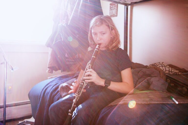 Jente sitter på seng og spiller klarinett. Ser ned. Ser litt trist ut.